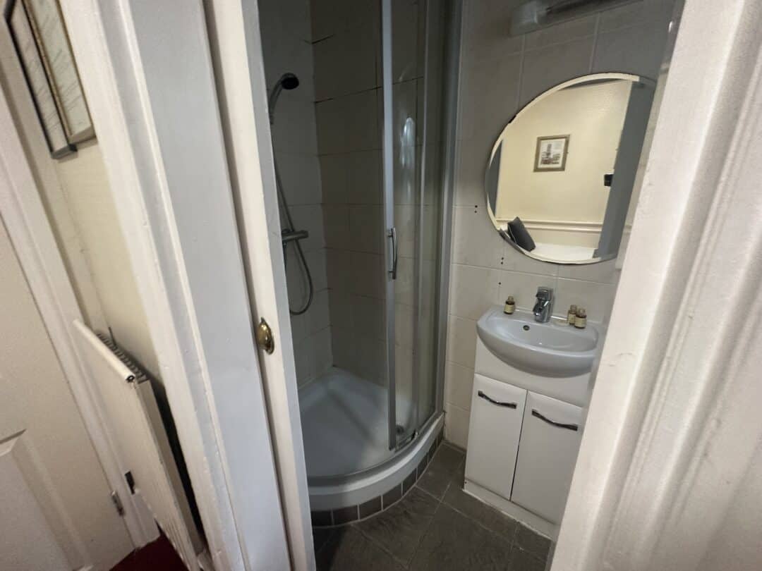 A small bathroom with a curved glass shower, a white sink cabinet, a round mirror above the sink, and bottles on the sink. A framed picture hangs on the wall, reflected in the mirror.