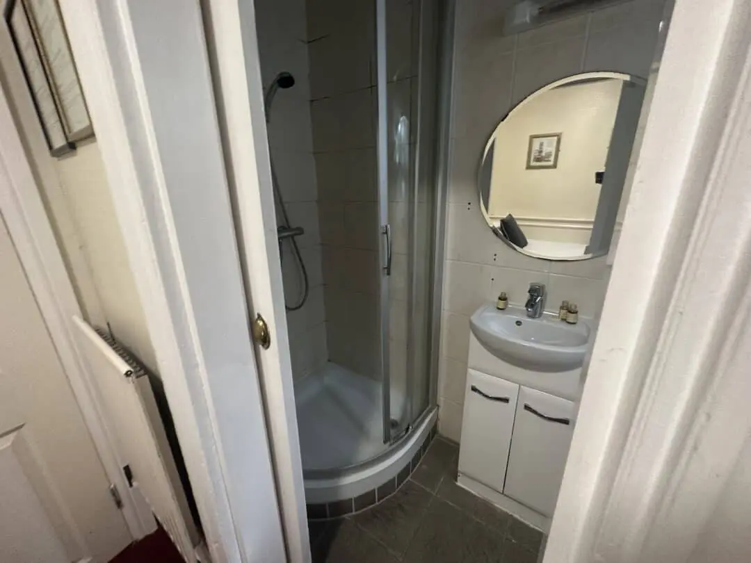 A small bathroom with a curved glass shower, a white sink cabinet, a round mirror above the sink, and bottles on the sink. A framed picture hangs on the wall, reflected in the mirror.