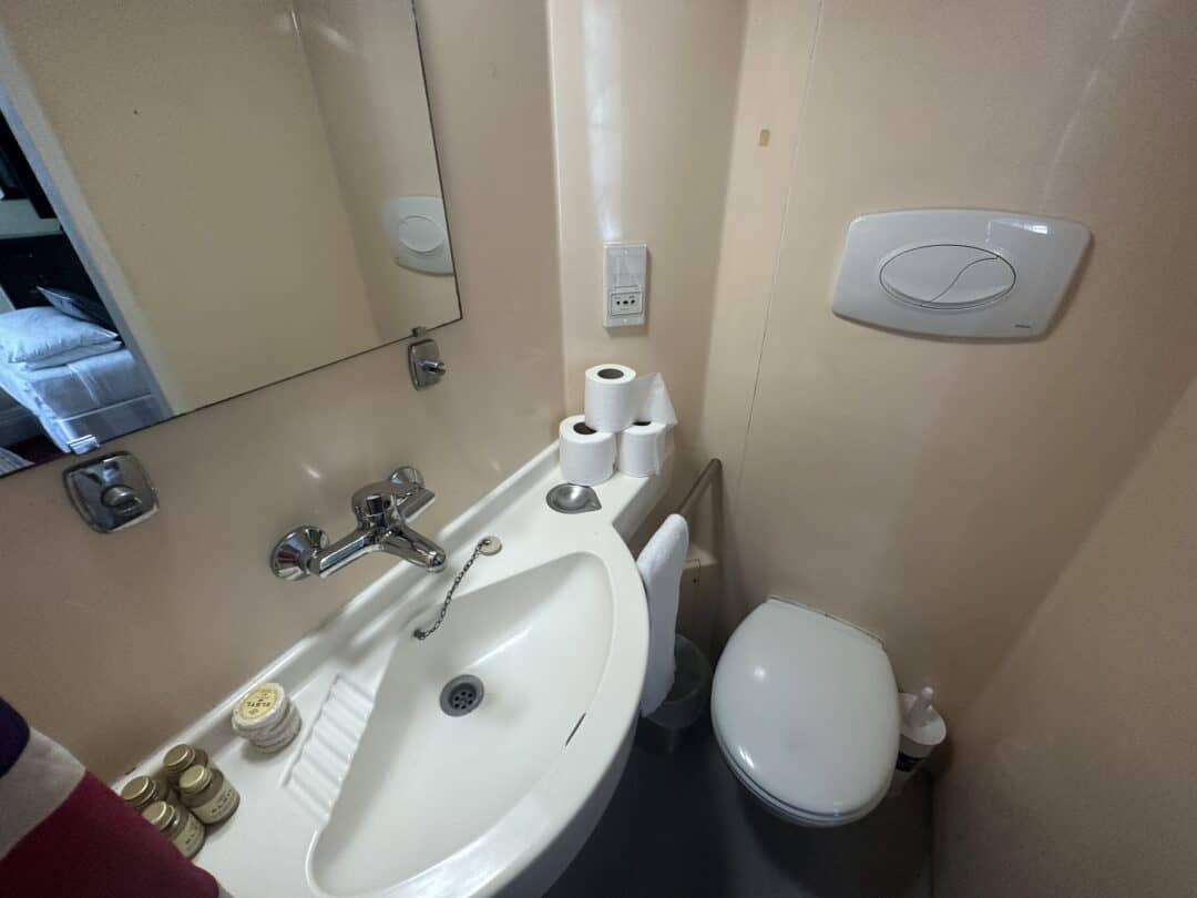 A small bathroom with a white sink, wall-mounted faucet, mirror, toilet, toilet paper rolls, and toiletries on the sink. The space is compact, with beige walls and gray flooring. A bed is partially visible in the mirror.