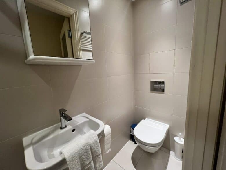 A small bathroom with beige tiles, featuring a wall-mounted sink with a towel, a mirror above it, and a wall-mounted toilet with a toilet paper holder and bin nearby.