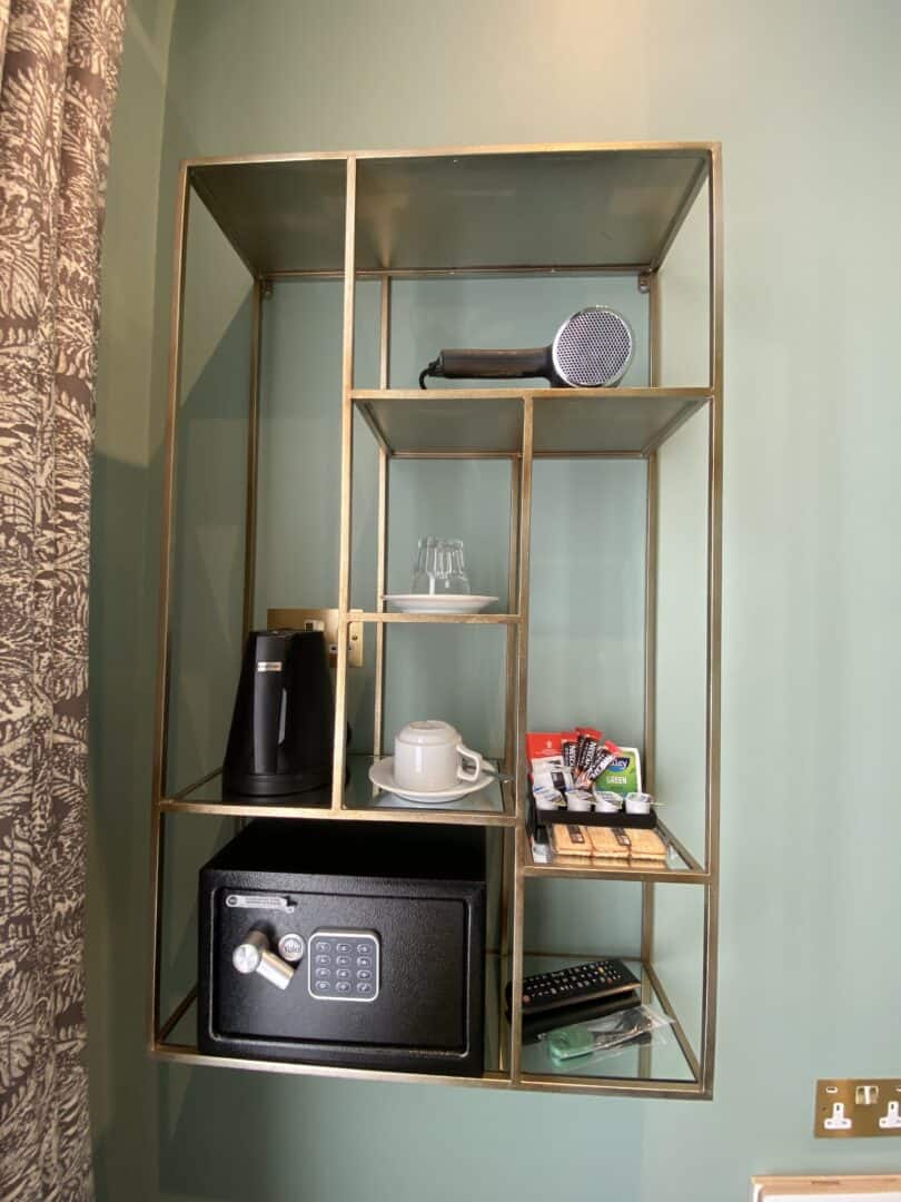 A gold-framed wall shelf holds a safe, electric kettle, cups, glasses, snacks, a hairdryer, TV remote, and an assortment of tea and coffee items against a green wall.