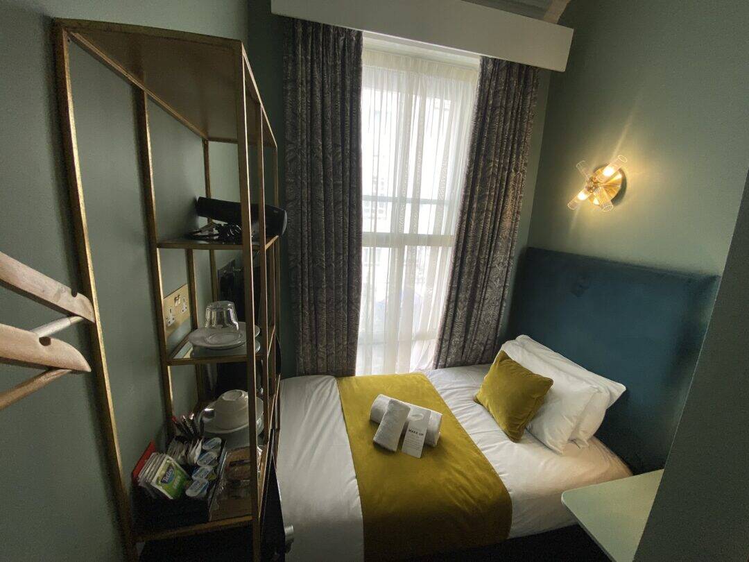 A small, cozy hotel room with a single bed, teal headboard, white sheets, a yellow throw and cushion. There’s a window with curtains, a shelving unit with amenities, and a wall lamp above the bed. A book lies open on the bed.