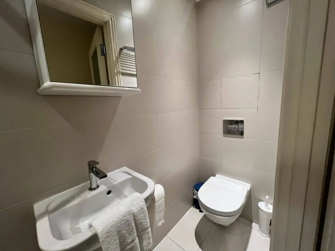 A small bathroom with beige tiles, featuring a wall-mounted sink with a towel, a mirror above it, and a wall-mounted toilet with a toilet paper holder and bin nearby.