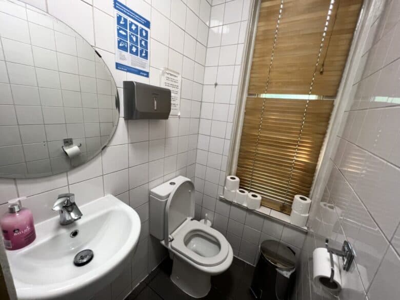 A small restroom with white tiled walls, a round mirror above a sink with pink soap, a toilet, several toilet rolls on a windowsill, a trash bin, a paper towel dispenser, and window blinds.