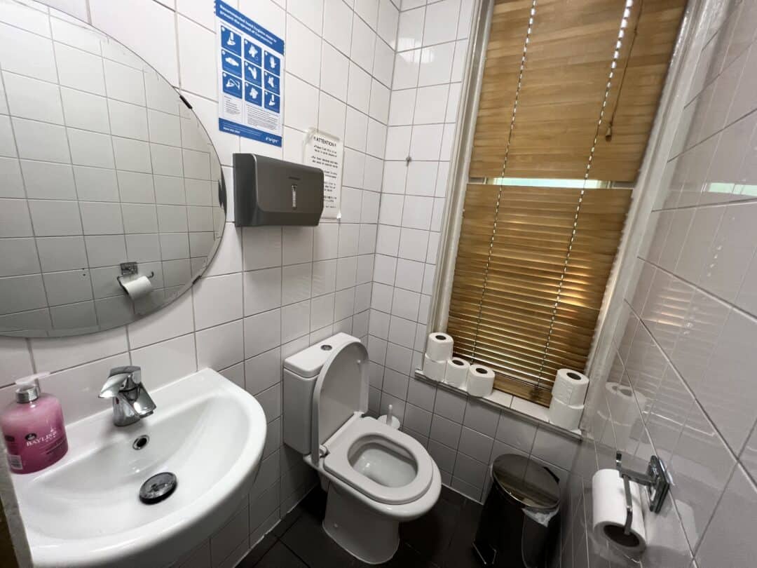 A small restroom with white tiled walls, a round mirror above a sink with pink soap, a toilet, several toilet rolls on a windowsill, a trash bin, a paper towel dispenser, and window blinds.