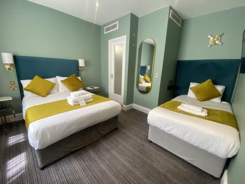 A hotel room with a double bed and a single bed, both with yellow throws and pillows, a set of folded towels on each bed, green walls, a wall mirror, and modern lighting.