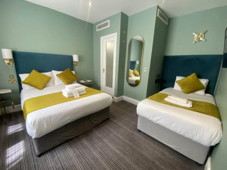 A hotel room with a double bed and a single bed, both with yellow throws and pillows, a set of folded towels on each bed, green walls, a wall mirror, and modern lighting.