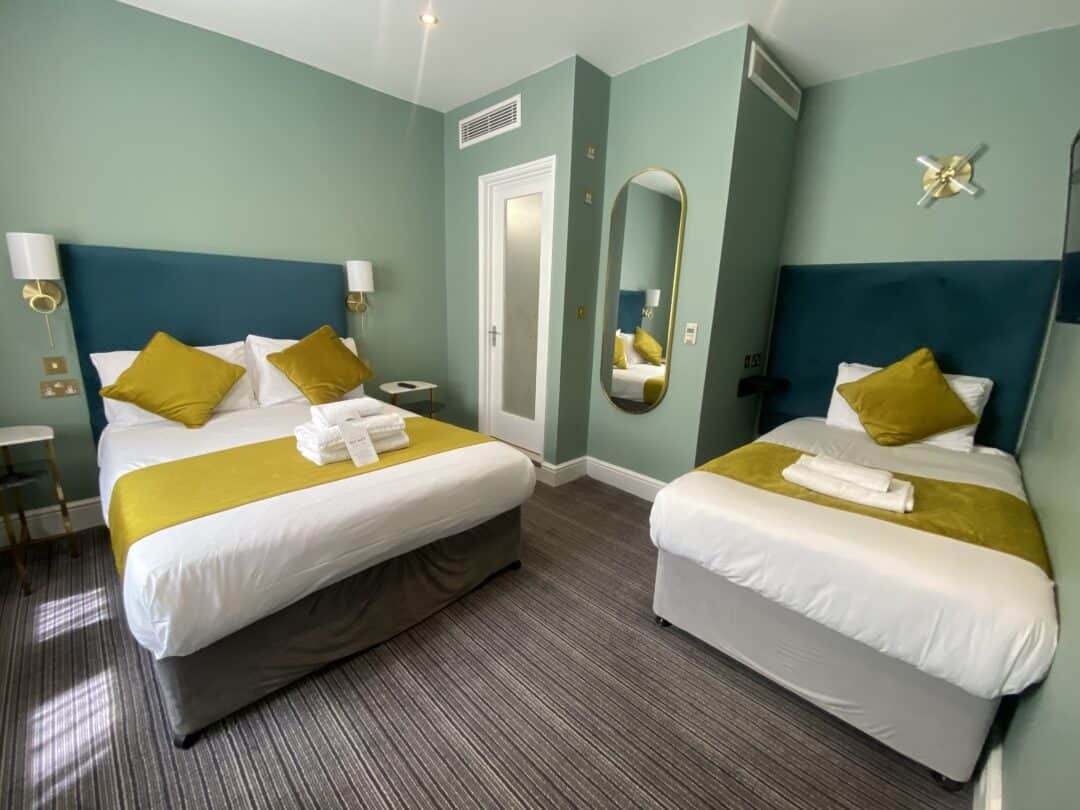 A hotel room with a double bed and a single bed, both with yellow throws and pillows, a set of folded towels on each bed, green walls, a wall mirror, and modern lighting.