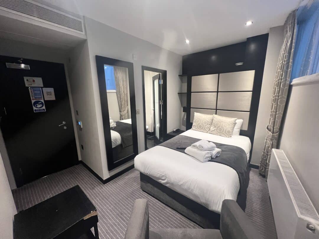 Modern hotel room with a neatly made double bed, gray and white decor, two mirrors on the wall, a window with curtains, and folded towels placed on the bed. A black door is visible in the background.