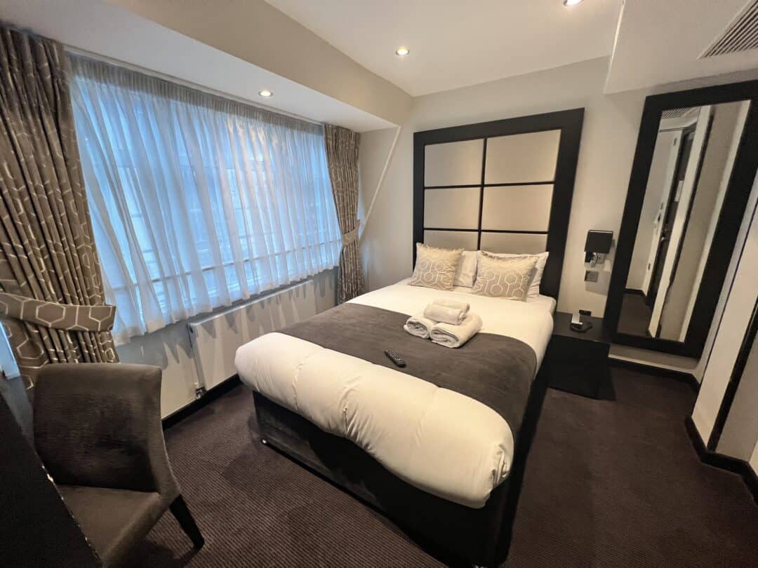 A modern hotel room with a neatly made double bed, two patterned pillows, folded towels, a remote, a chair by the window with curtains, and a floor-length mirror on the wall beside the bed.