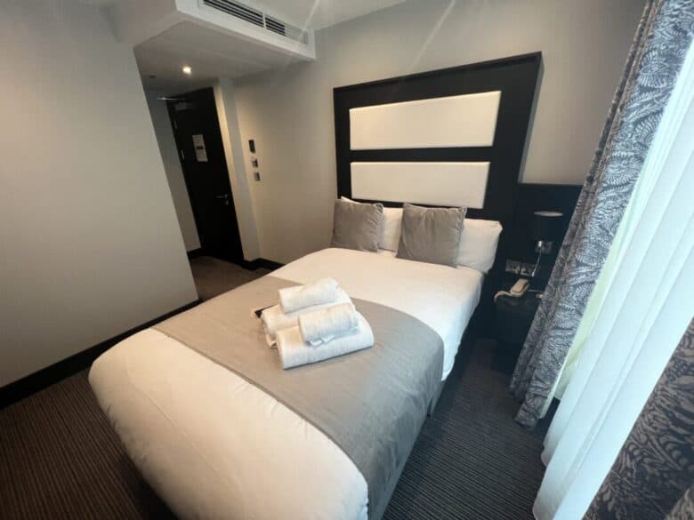 A neatly made bed with white and gray bedding, two gray pillows, and folded white towels is in a modern hotel room with neutral walls, soft lighting, and a window with patterned curtains.