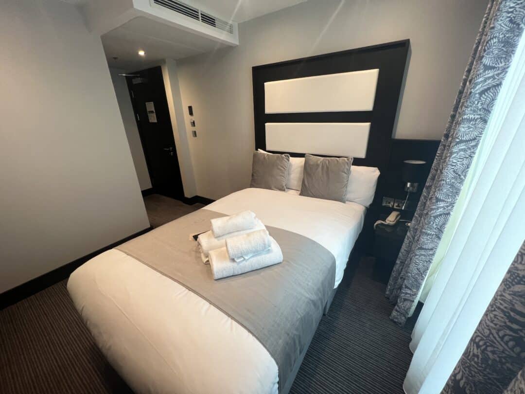 A neatly made bed with white and gray bedding, two gray pillows, and folded white towels is in a modern hotel room with neutral walls, soft lighting, and a window with patterned curtains.