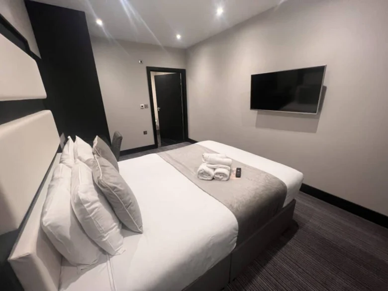 Modern hotel room with a neatly made double bed, folded towels and a remote control on top, a wall-mounted TV, and a door in the background; decorated in neutral tones with soft lighting.