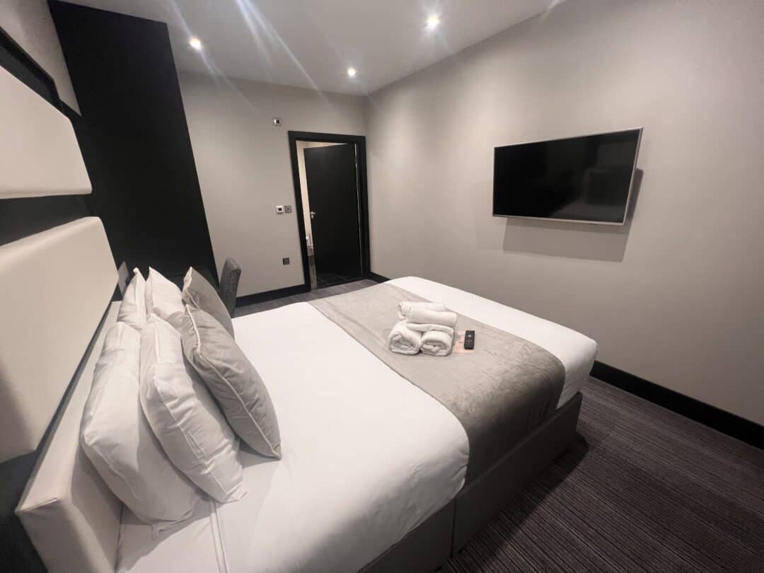 Modern hotel room with a neatly made double bed, folded towels and a remote control on top, a wall-mounted TV, and a door in the background; decorated in neutral tones with soft lighting.