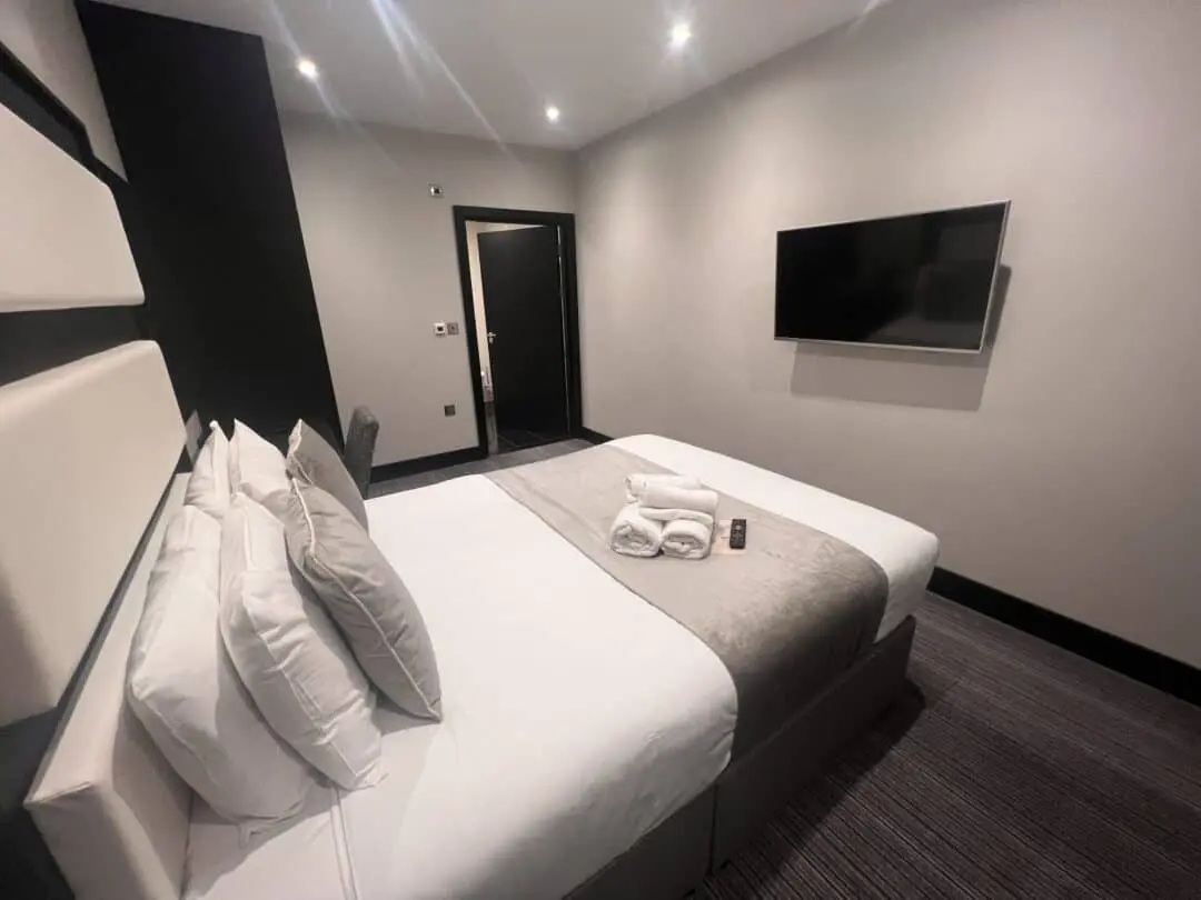 Modern hotel room with a neatly made double bed, folded towels and a remote control on top, a wall-mounted TV, and a door in the background; decorated in neutral tones with soft lighting.