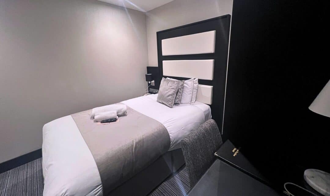 A modern hotel room with a single bed dressed in white and gray bedding, with neatly folded towels and a TV remote on top. There is a padded headboard, a chair, a desk, and neutral-colored walls and carpet.