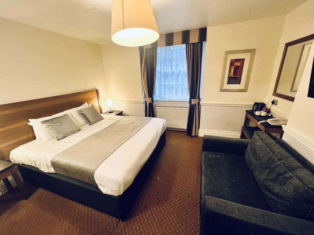 A hotel room with a double bed, gray couch, bedside tables with lamps, a window with striped curtains, a wall mirror, a framed picture, and a corner desk with a kettle and cups. The carpet is brown.