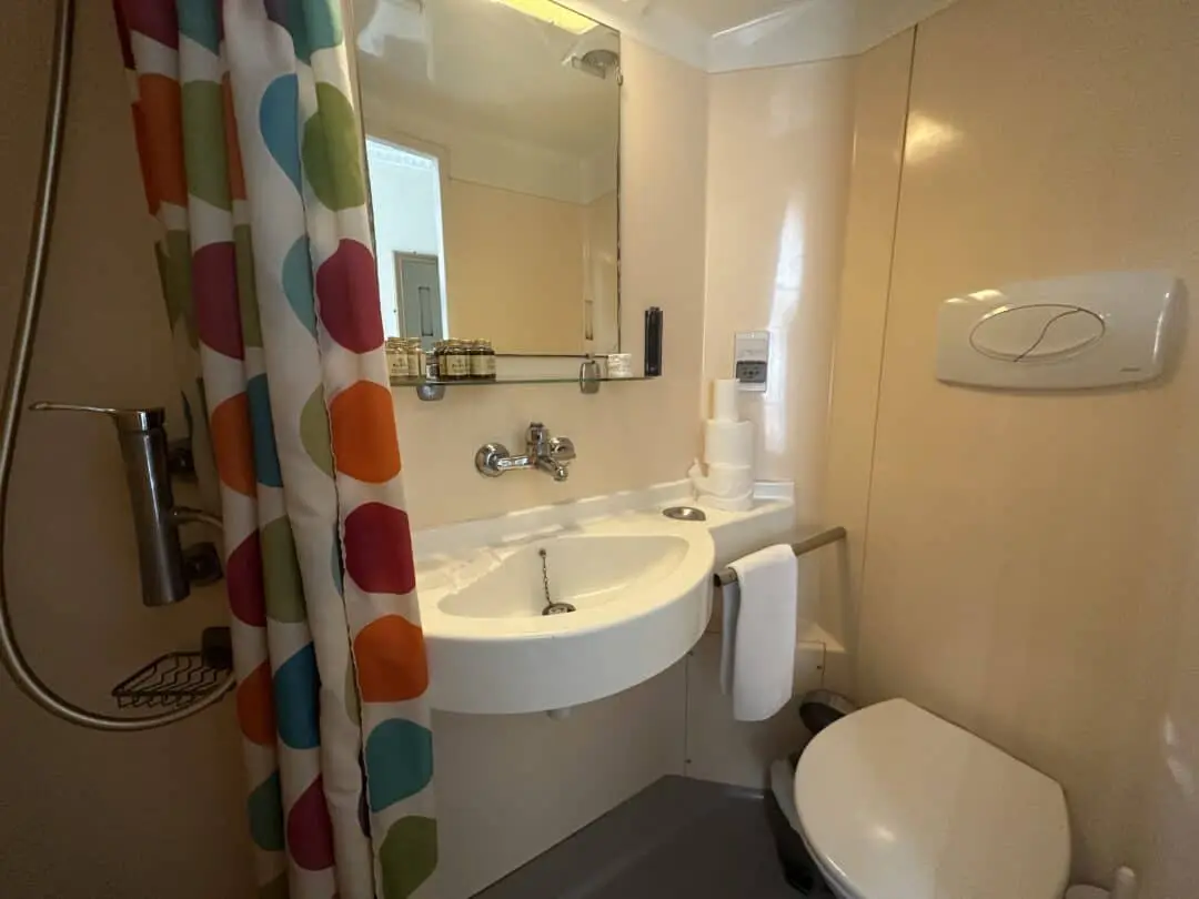 A compact bathroom with a colorful polka dot shower curtain, a wall-mounted sink with a mirror, toiletries, toilet paper rolls, a towel, and a toilet, all in a beige-toned space.