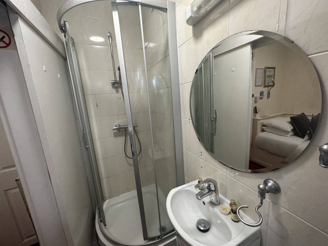A compact bathroom with a corner shower cabin, a small white sink, a round mirror above the sink, and toiletries on the sink. The reflection in the mirror shows a bed and wall decor in the adjacent room.