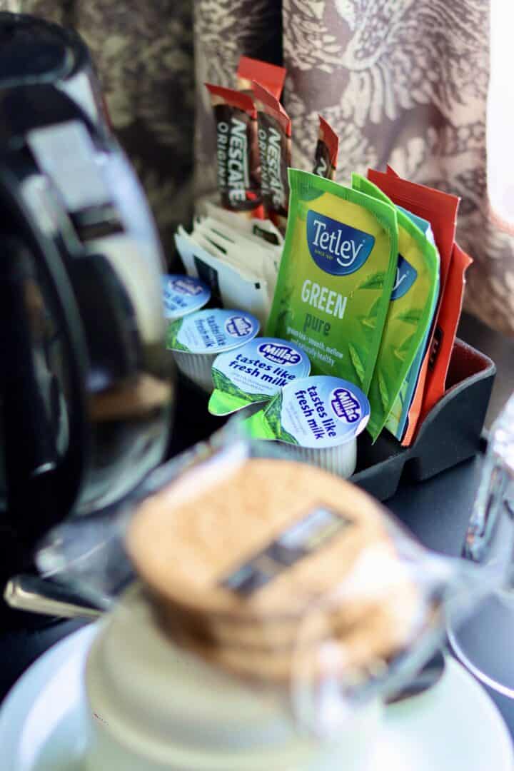 A close-up of a beverage station with Tetley green tea bags, Nescafé coffee sticks, milk pods, sugar packets, and biscuits, set beside a coffee maker and a patterned curtain in the background.