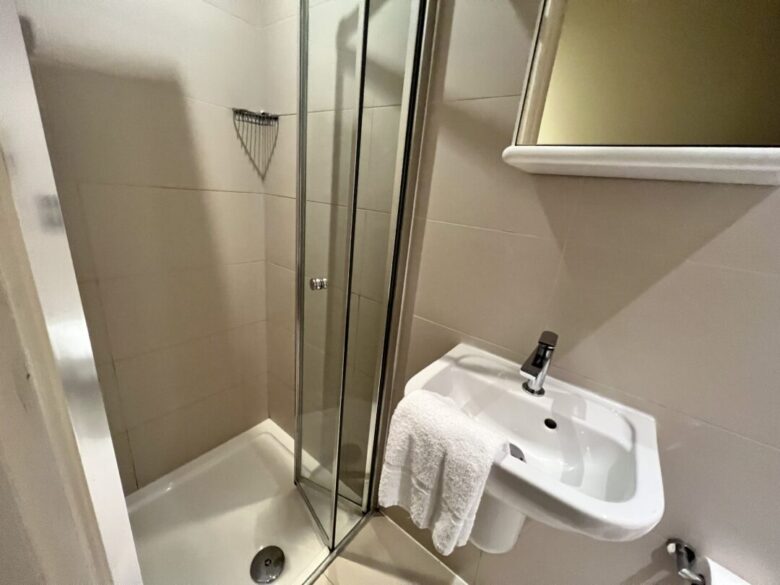 Small bathroom with a glass-enclosed shower, a white sink with a chrome faucet, a white towel draped over the sink, and a rectangular mirror above the sink. The walls are light beige tiles.