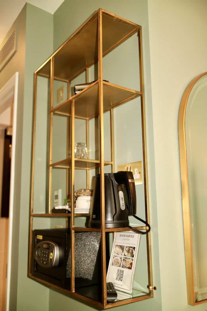 A gold-framed wall shelf holds a black electric kettle, a safe, glasses, a menu, and other small items against a light green wall in a room.