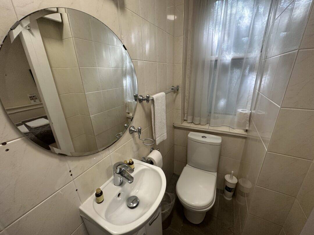 Small bathroom with a round mirror above a white sink, two small bottles on the sink, a towel on a rack above the toilet, frosted window with sheer curtains, and a toilet paper holder on the windowsill.