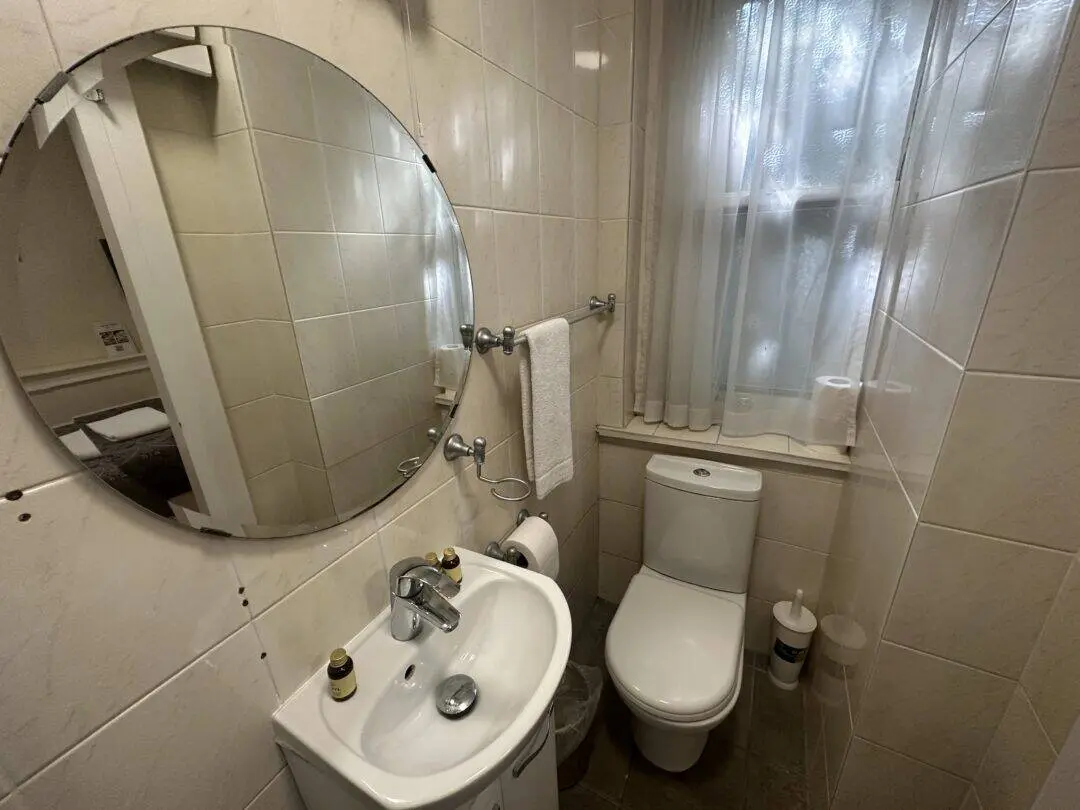Small bathroom with a round mirror above a white sink, two small bottles on the sink, a towel on a rack above the toilet, frosted window with sheer curtains, and a toilet paper holder on the windowsill.