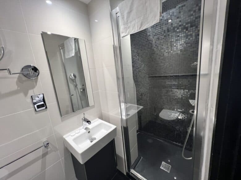 Modern bathroom with white tiles, a wall-mounted sink, a mirror, and a glass-enclosed shower with dark mosaic tiles. A white towel hangs above the shower, and a hairdryer is visible next to the mirror.