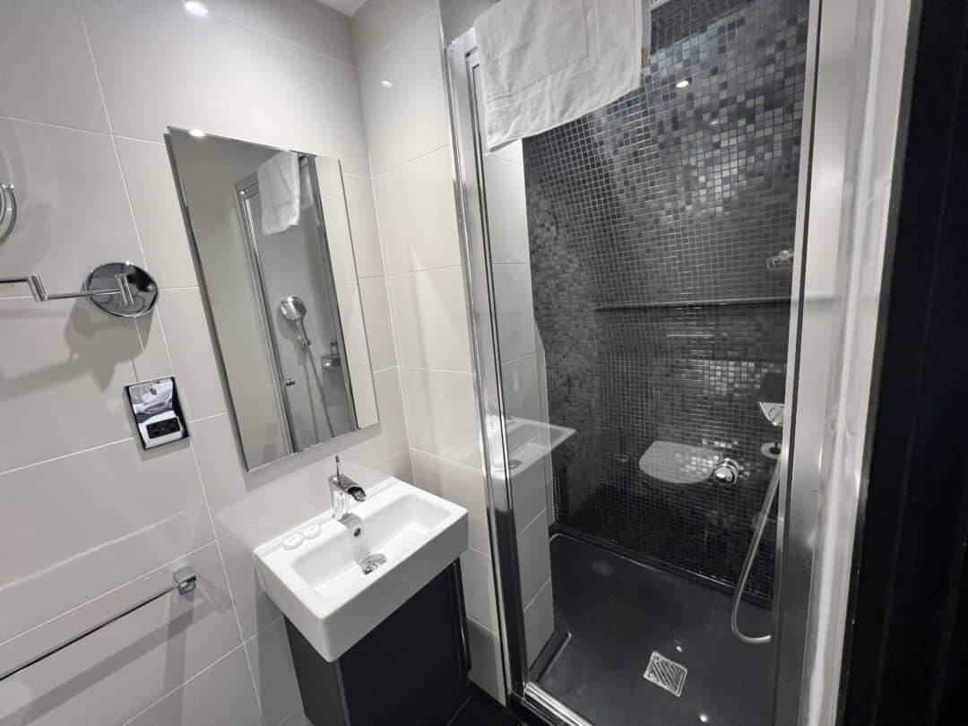Modern bathroom with white tiles, a wall-mounted sink, a mirror, and a glass-enclosed shower with dark mosaic tiles. A white towel hangs above the shower, and a hairdryer is visible next to the mirror.