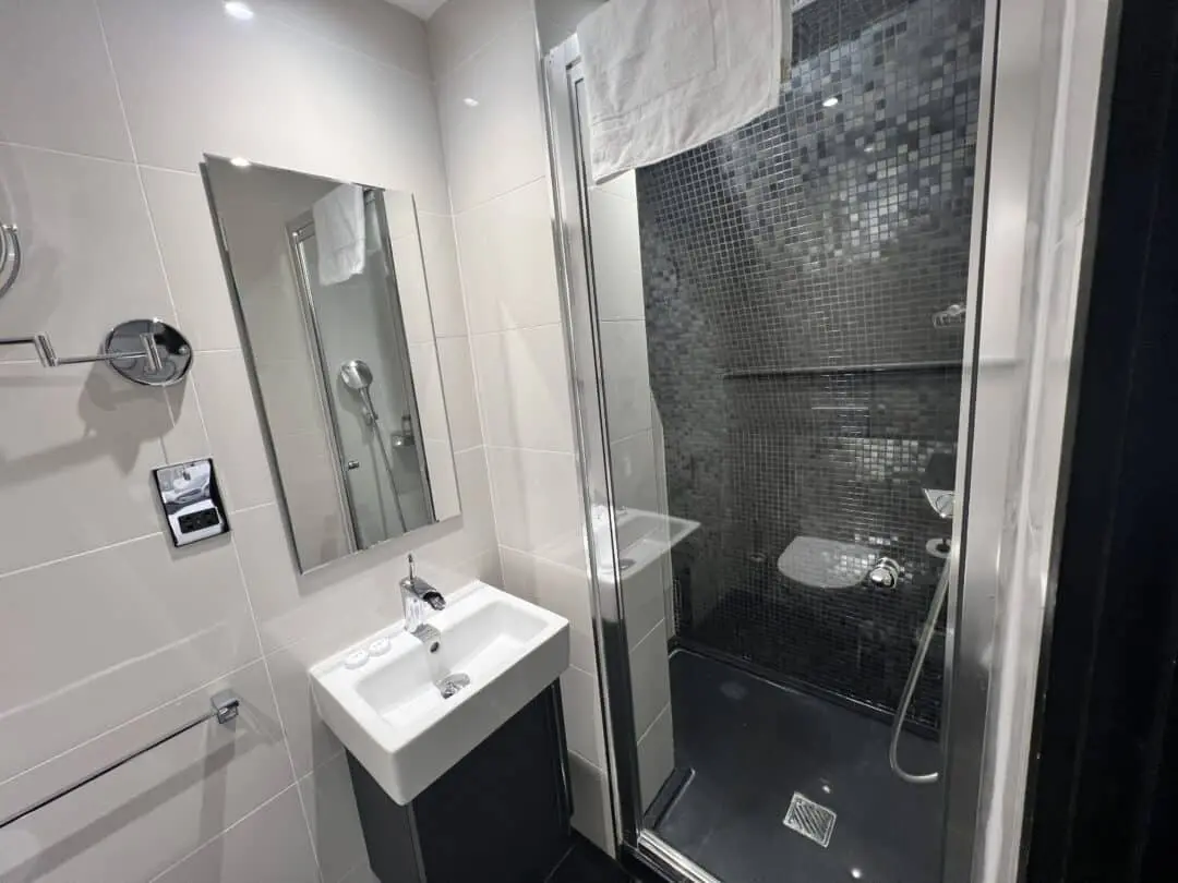 Modern bathroom with white tiles, a wall-mounted sink, a mirror, and a glass-enclosed shower with dark mosaic tiles. A white towel hangs above the shower, and a hairdryer is visible next to the mirror.