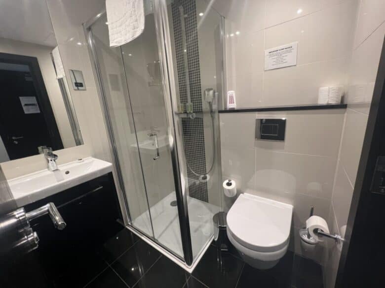 Modern bathroom with a glass shower enclosure, wall-mounted toilet, white sink, and black tile floor. Towels, toilet paper, and toiletries are visible, along with a mirror reflecting the room.
