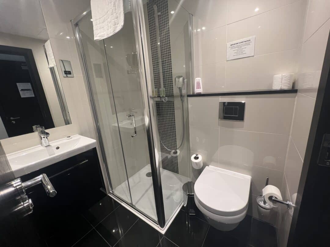 Modern bathroom with a glass shower enclosure, wall-mounted toilet, white sink, and black tile floor. Towels, toilet paper, and toiletries are visible, along with a mirror reflecting the room.