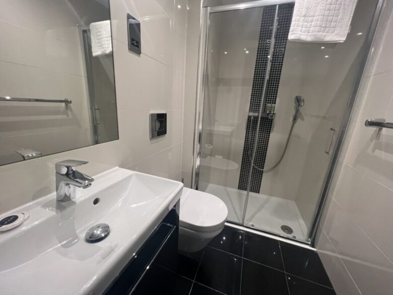 Modern bathroom with white sink, wall-mounted toilet, and glass-enclosed shower with a handheld showerhead. The room has black floor tiles and a white towel hanging above the shower.