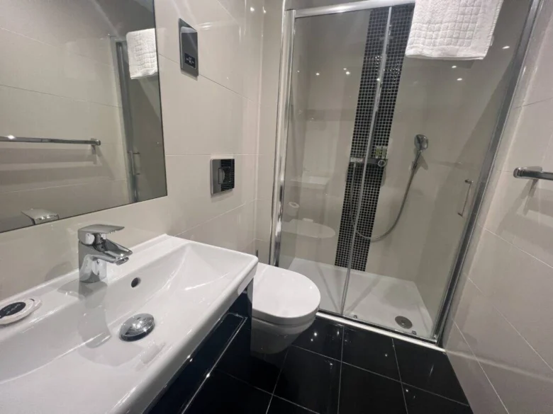 Modern bathroom with white sink, wall-mounted toilet, and glass-enclosed shower with a handheld showerhead. The room has black floor tiles and a white towel hanging above the shower.