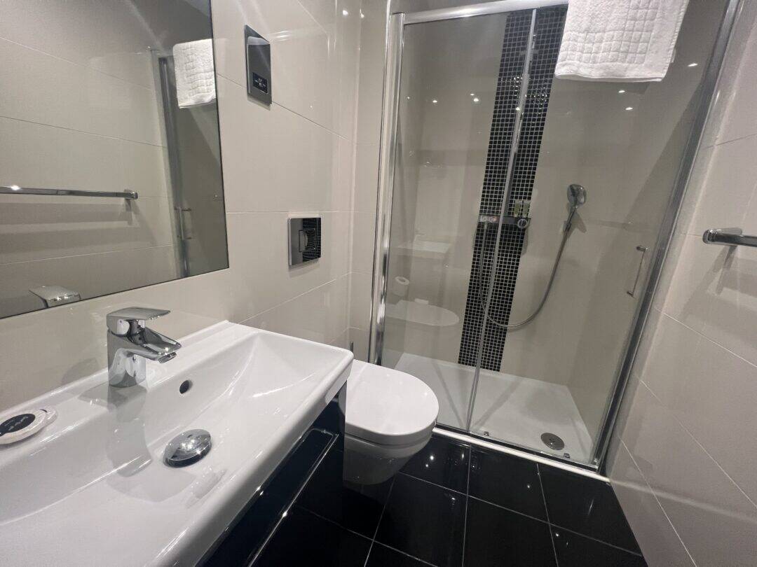 Modern bathroom with white sink, wall-mounted toilet, and glass-enclosed shower with a handheld showerhead. The room has black floor tiles and a white towel hanging above the shower.