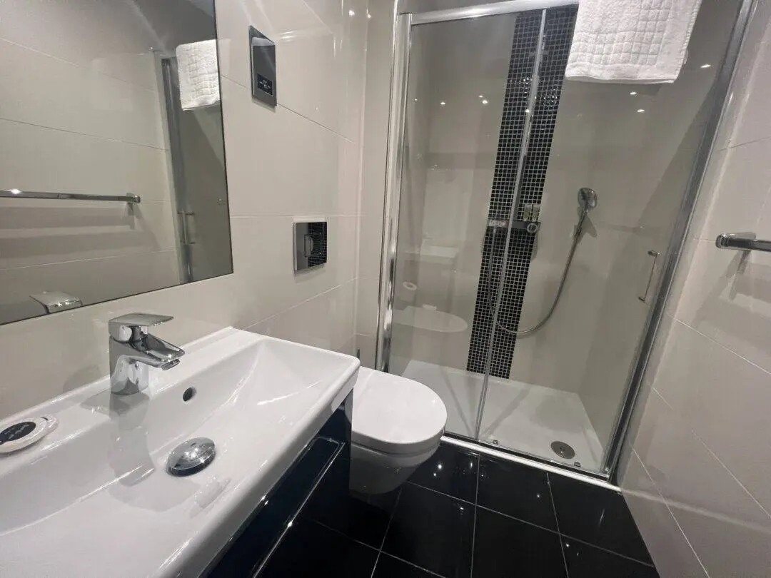 Modern bathroom with white sink, wall-mounted toilet, and glass-enclosed shower with a handheld showerhead. The room has black floor tiles and a white towel hanging above the shower.