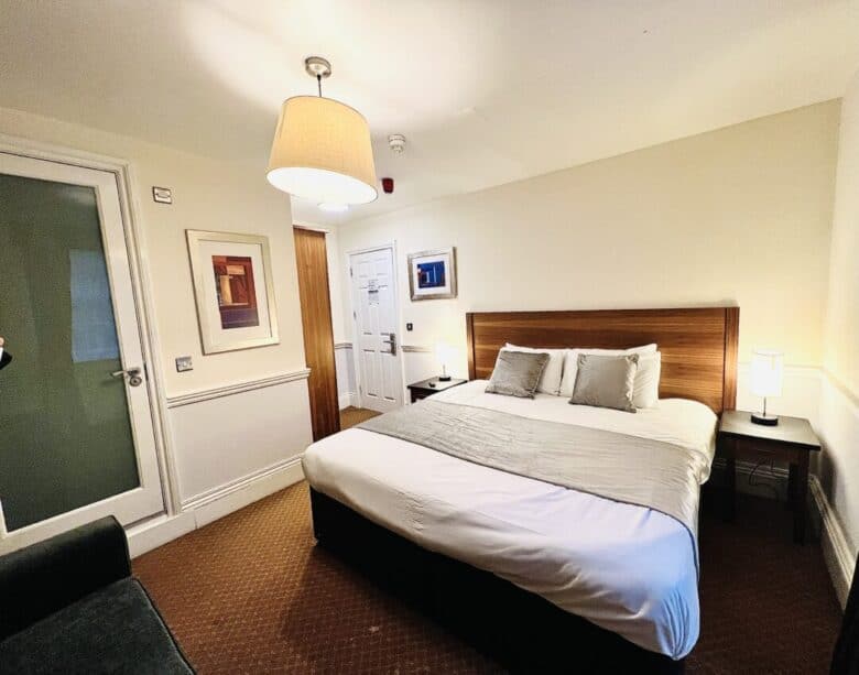 A cozy hotel room with a large bed, two bedside tables with lamps, framed artwork on the walls, a green sofa, and a closed door. The lighting is warm and inviting.
