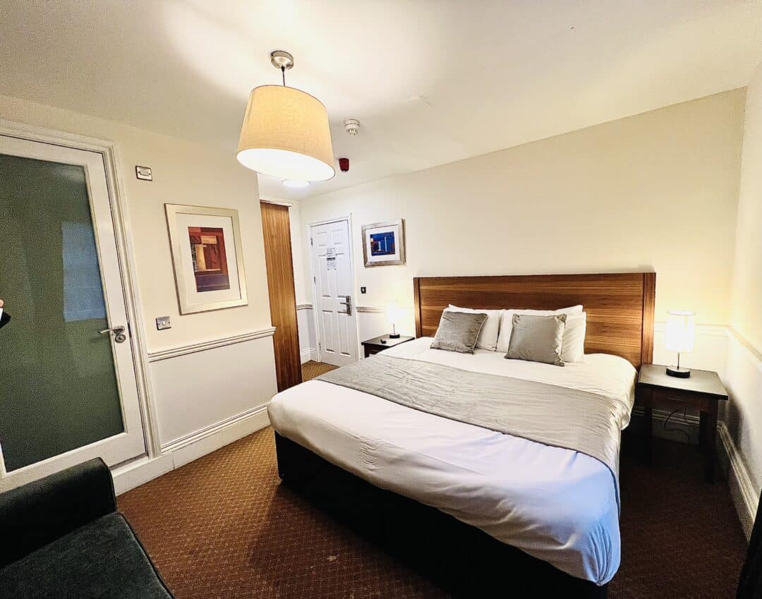 A cozy hotel room with a large bed, two bedside tables with lamps, framed artwork on the walls, a green sofa, and a closed door. The lighting is warm and inviting.