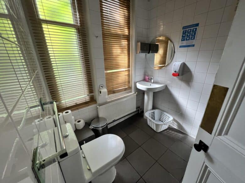 A clean bathroom with white tiled walls, a toilet, sink, mirror, soap dispenser, laundry basket, and large windows with wooden blinds letting in natural light. Toilet paper rolls and a radiator are also visible.