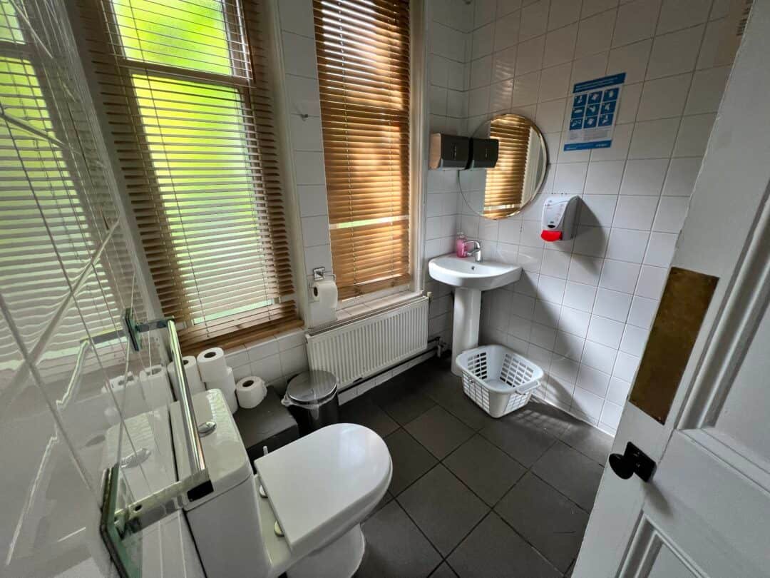 A clean, tiled bathroom with a toilet, sink, mirror, window with wooden blinds, a laundry basket, and toilet paper rolls on a shelf above the radiator. Natural light comes through the blinds.