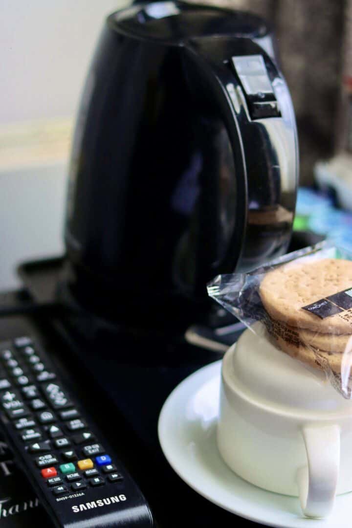A black electric kettle, a TV remote, and a white cup with a saucer holding packaged biscuits are arranged on a dark surface.