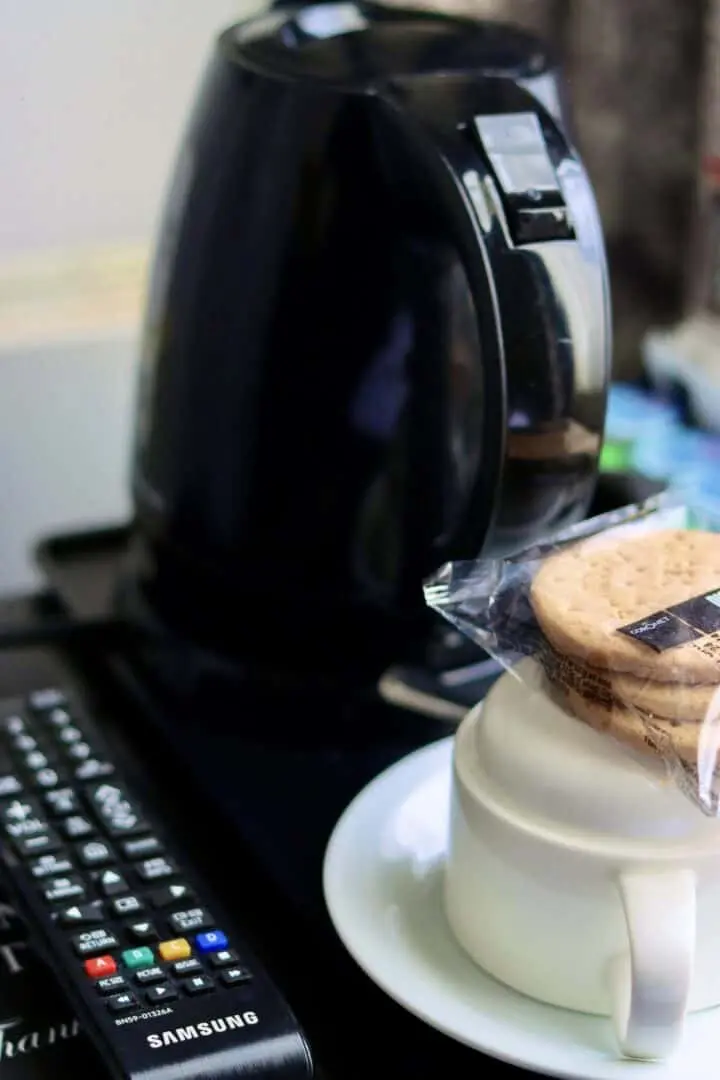A black electric kettle sits behind a white cup and saucer with packaged biscuits on top. A TV remote control is visible in the foreground.