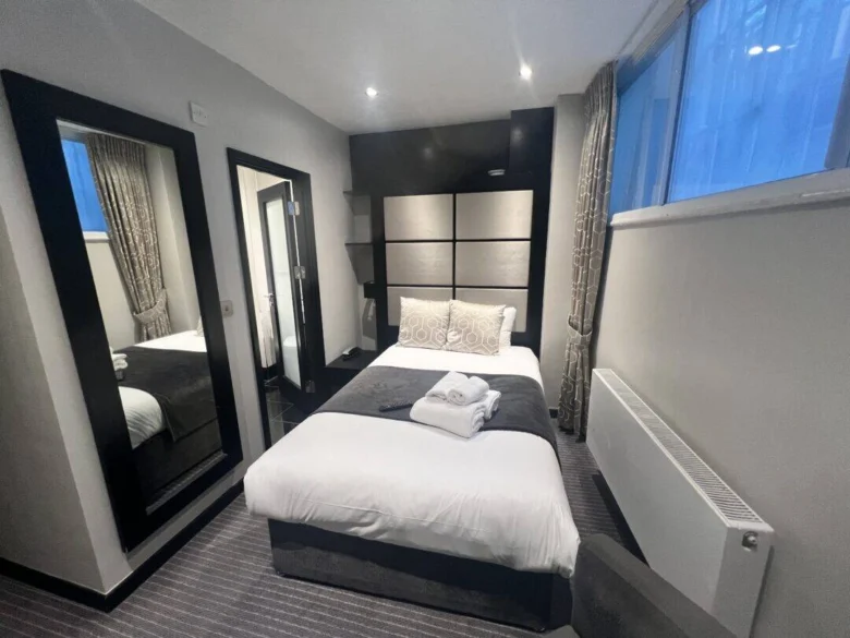 A modern, compact hotel room with a single bed, folded towels on top, a large mirror, window with curtains, radiator, and a door leading to a bathroom. The decor is neutral with gray and white tones.