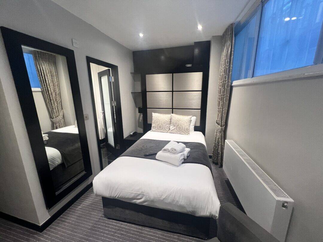 A modern, compact hotel room with a single bed, folded towels on top, a large mirror, window with curtains, radiator, and a door leading to a bathroom. The decor is neutral with gray and white tones.