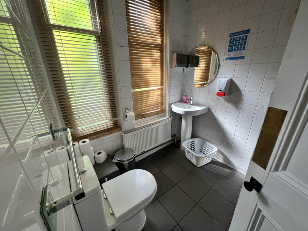 A clean, tiled bathroom with a toilet, sink, round mirror, soap dispenser, several toilet paper rolls, a white laundry basket, and large windows with wooden blinds letting in natural light.