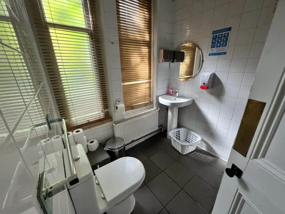 A clean, tiled bathroom with a toilet, sink, round mirror, soap dispenser, several toilet paper rolls, a white laundry basket, and large windows with wooden blinds letting in natural light.