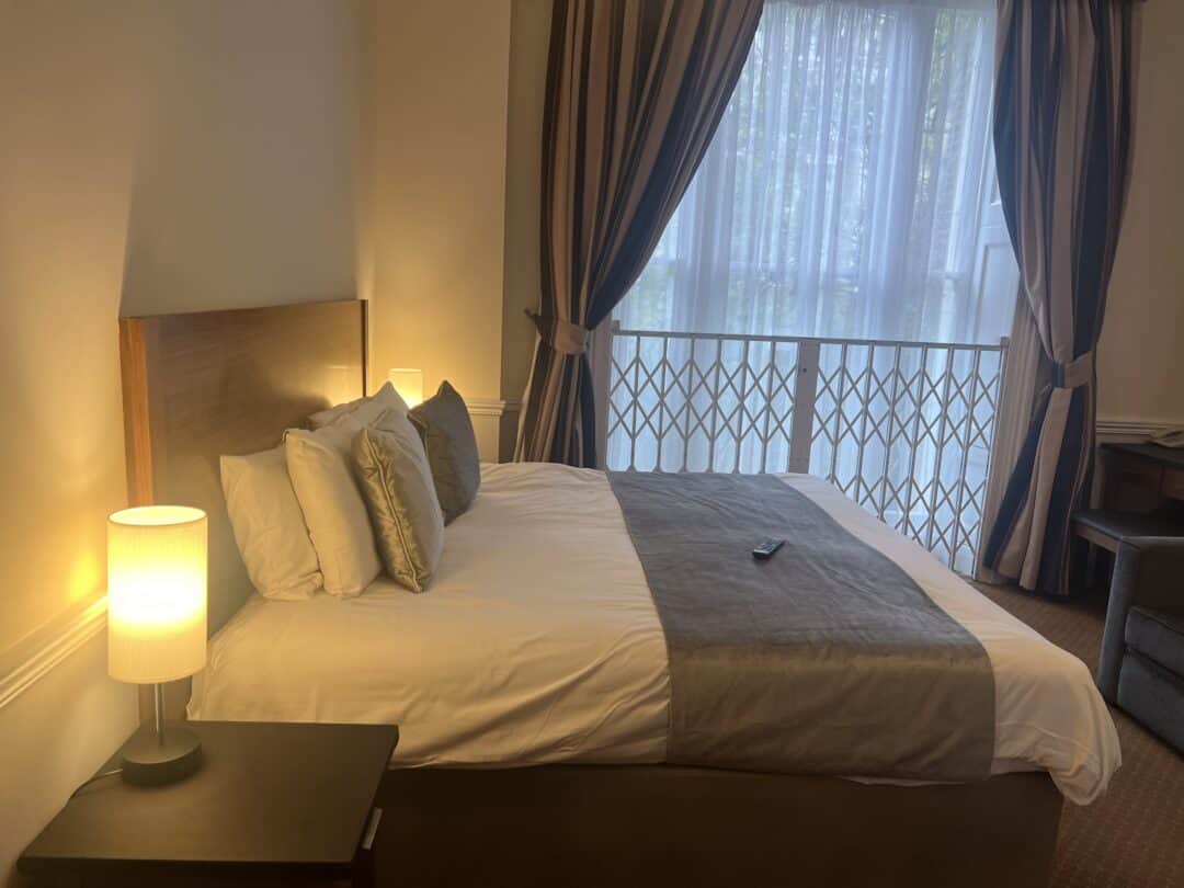 A neatly made bed with white and gray bedding and multiple pillows is next to a lit bedside lamp. Large windows with sheer curtains and drapes let in natural light, and a TV remote rests on the bed.