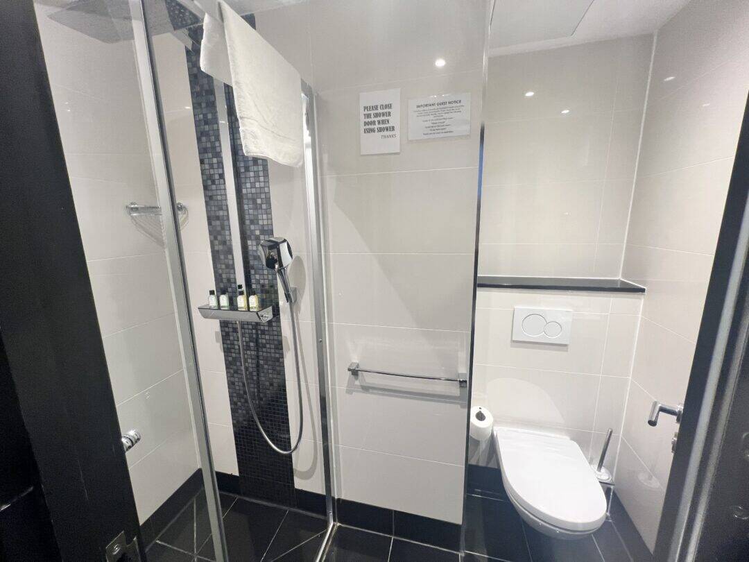 Modern bathroom with a glass-enclosed shower, towel, toiletries on a shelf, and a wall-mounted toilet. Beige tiles and dark accents give a clean, contemporary look. A sign is posted on the wall above the towel rack.