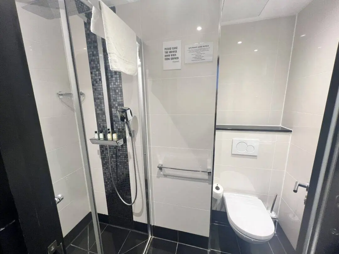 Modern bathroom with a glass-enclosed shower, towel, toiletries on a shelf, and a wall-mounted toilet. Beige tiles and dark accents give a clean, contemporary look. A sign is posted on the wall above the towel rack.